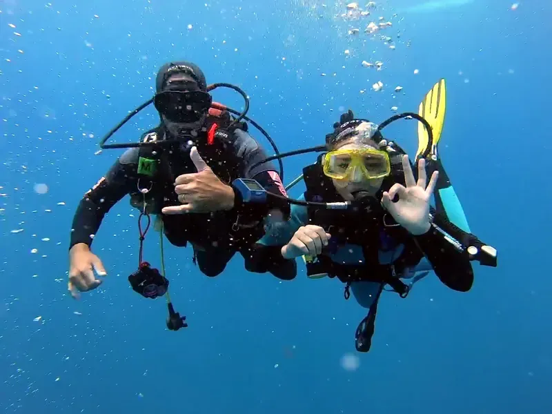 Raíssa Felipetto, divemaster da Atlantes, e o instrutor da equipe em mergulho de naufrágio, fazendo sinais de OK em água azul